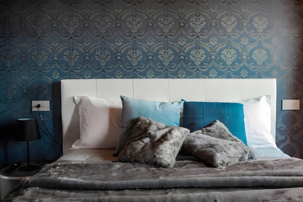 Quarto com papel de parede estampado, cabeceira branca e cama com várias almofadas.