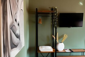 Een kamer met een groot abstract schilderij, een aan de muur bevestigde televisie, een houten plank met een vaas en decoratieve objecten, en een bed met een witte deken.