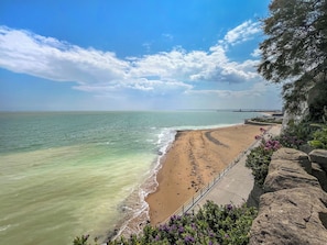 Ein Küstenstrand mit blauem Himmel, sandigem Ufer und einem felsigen Klippenhang mit Vegetation.