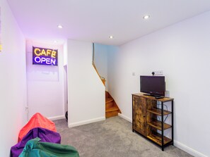 Une pièce avec un escalier, un téléviseur posé sur un meuble en bois et un panneau néon indiquant que le café est ouvert.