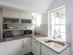 A kitchen with white cabinets, a microwave, a toaster, a sink, and a window with a view of greenery. - Old Anchor Cottage - UK51953 (Broadstairs)
