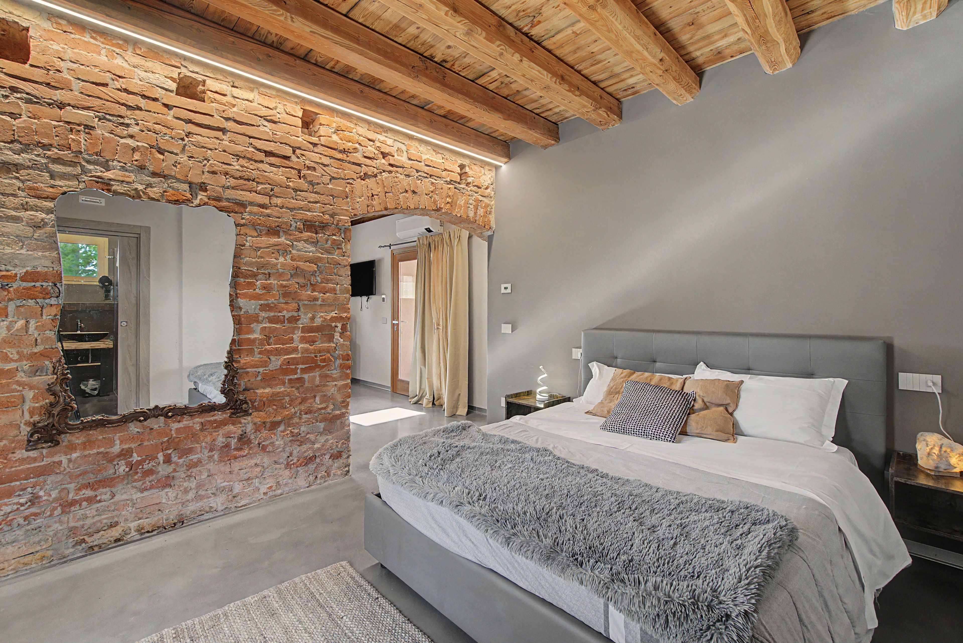 Une chambre avec un mur en briques, un grand lit avec de nombreux oreillers, une tête de lit grise et une lampe de chevet moderne.