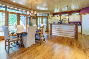 A spacious kitchen with wooden cabinets and a central island, large windows offering a view, and a dining area with a table and chairs. - Log Home on Tipsico Lake w/ Beach & Dock (Holly)