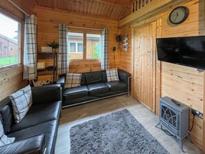 A cozy cabin interior with a black leather sofa, a grey rug, a television, and a wood stove. - Log Cabin Three - UK51961 (Sutton-on-Sea)