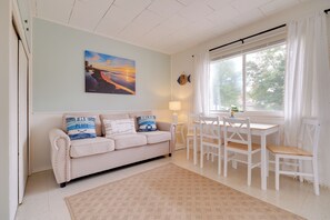 A cozy living room with a beige sofa, a dining table with chairs, a painting of a sunset, and a window with curtains.