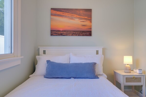 A bedroom with a bed, pillows, a nightstand with a lamp, and a wall-mounted picture of a sunset over the ocean.