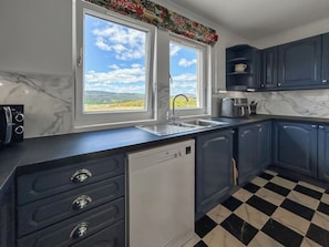 A kitchen with a checkered floor, a dishwasher, a sink, and a window with a view of a landscape. - Roanbank - The Hidden Road Retreat (Castle Douglas)