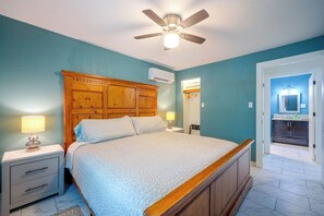 Ein Schlafzimmer mit einem hölzernen Kopfteil, einem Deckenventilator, einem Bett mit gemusterten Betttextilien, Nachttischen mit Lampen und einem durch eine offene Tür einsehbaren Badezimmer.