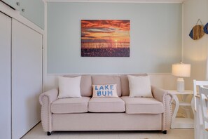 A beige sofa with 'LAKE BUM' pillows, a wall-mounted sunset painting, and a white side table with a lamp. - Beach Access! Sunrise Side Resort Retreat (Greenbush)