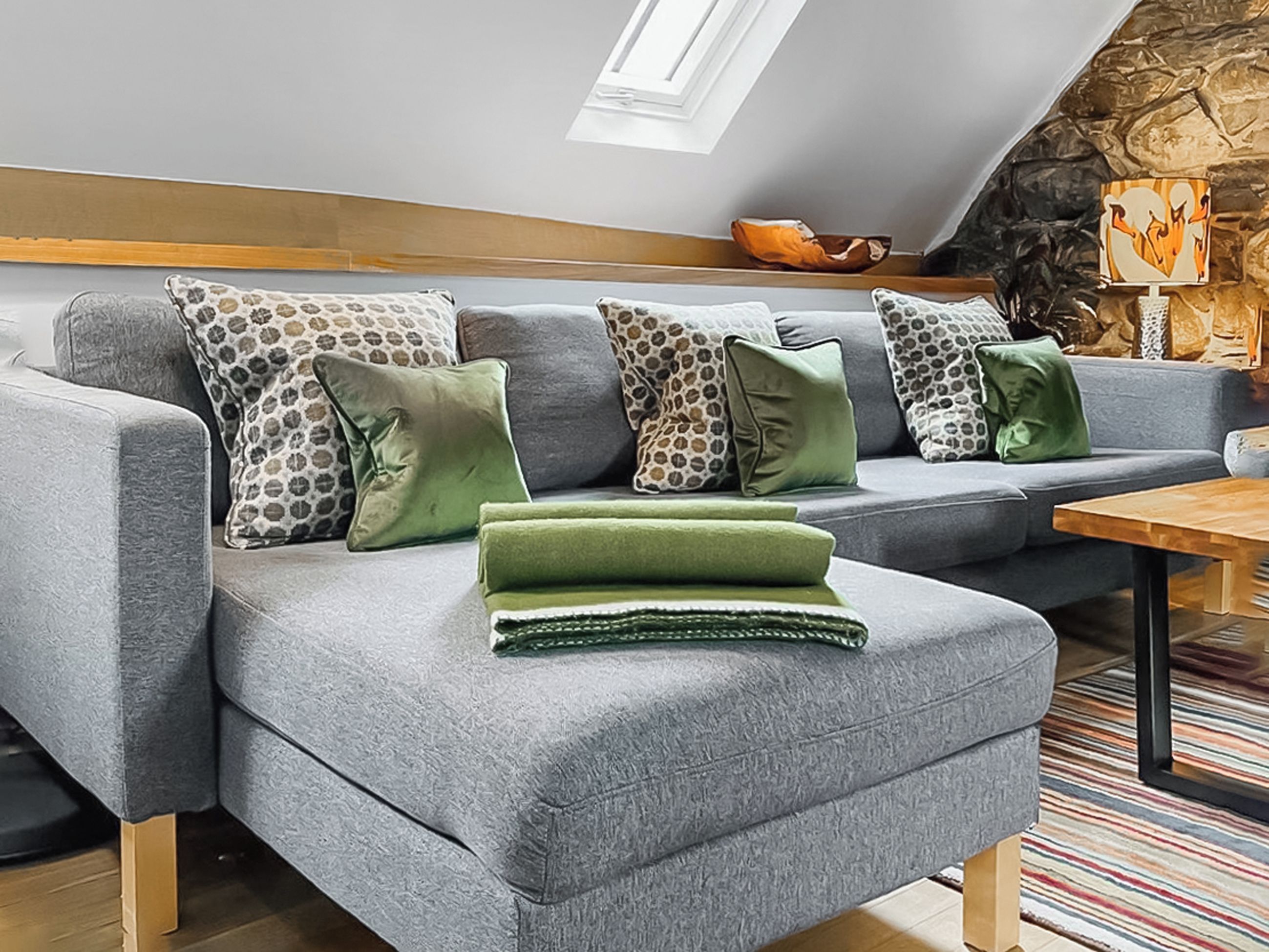 A grey sectional sofa with patterned and solid green cushions, a wooden coffee table, and a stone wall with a fireplace.