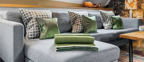 A grey sectional sofa with patterned and solid green cushions, a wooden coffee table, and a stone wall with a fireplace.