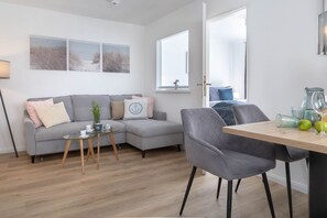 A modern living room with a grey sofa, a glass coffee table, and a dining area with a wooden table and grey chairs. - Marien4 - Möve1 - Prime Location - Right on the Beach (Sylt)