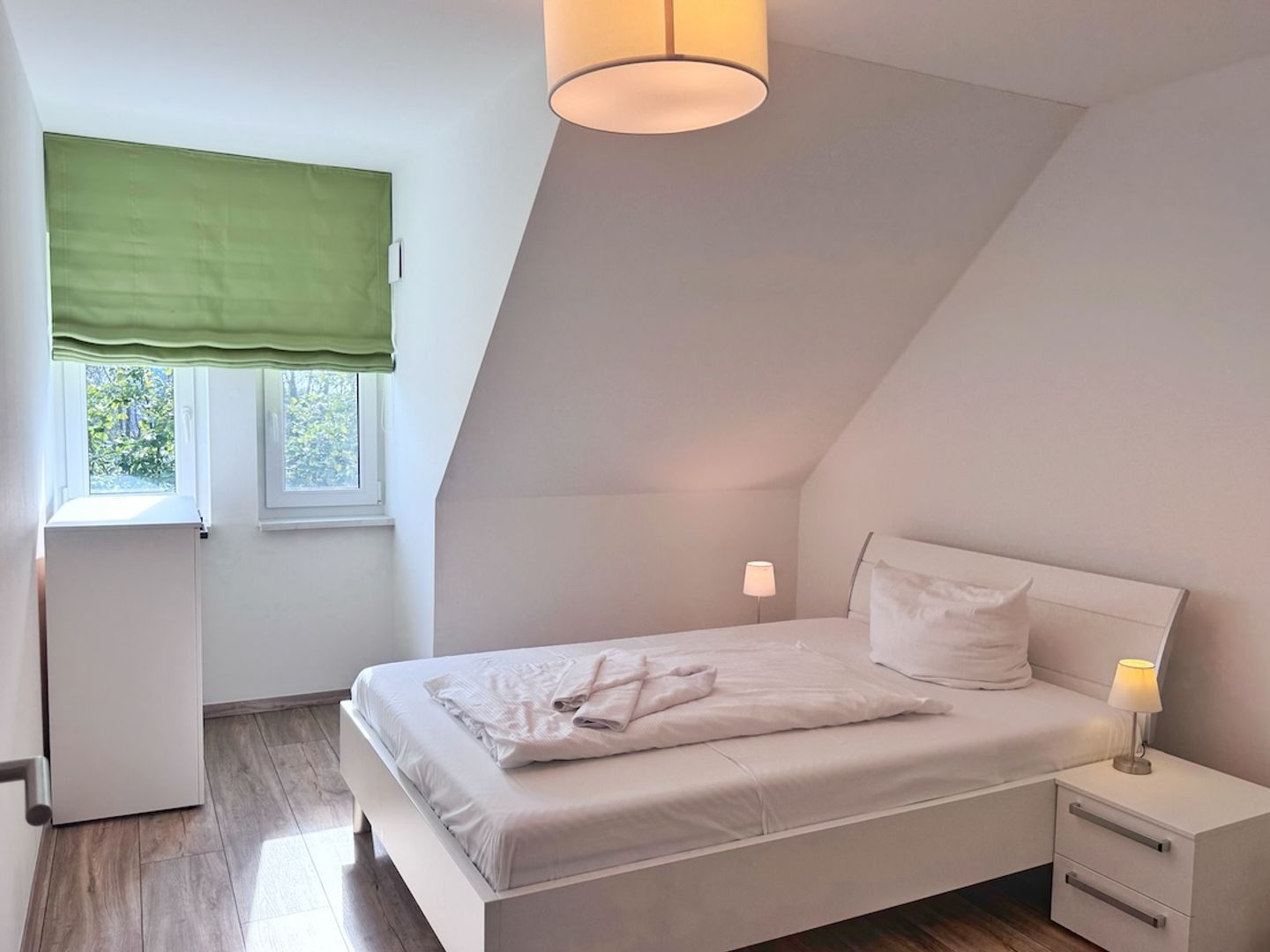 A bedroom with a bed, bedside tables, a window with a green blind, and a ceiling light.
