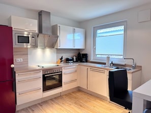 A compact kitchen with a microwave, oven, and a window with blinds. - Modern three-bedroom semi-detached home with a fireplace - Mein Glowe - DHH Links (Glowe)