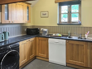 A compact kitchen with wooden cabinets, a washing machine, a dishwasher, a microwave, and a window with a view of greenery. - 2 bedroom accommodation in Holsworthy (Holsworthy)