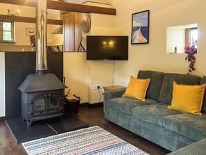 A living room with a wood-burning stove, a sofa with yellow cushions, a television mounted on the wall, and a patterned rug on the floor. - 2 bedroom accommodation in Holsworthy (Holsworthy)