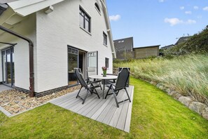 Une maison blanche avec une terrasse, une table entourée de chaises et un jardin avec de la pelouse et des rochers.