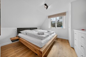 A bedroom with a wooden bed, white bedding, a bedside lamp, a window with a view, and a white dresser. - Senhüs - Stylish cottage for 6 people with a sauna, garden, and free E (Hörnum (Sylt))