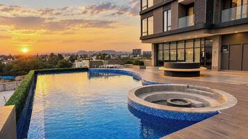 A rooftop swimming pool with a modern design, surrounded by a building with large windows and a view of the sunset.