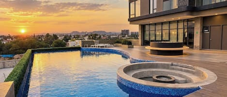 A rooftop swimming pool with a modern design, surrounded by a building with large windows and a view of the sunset.