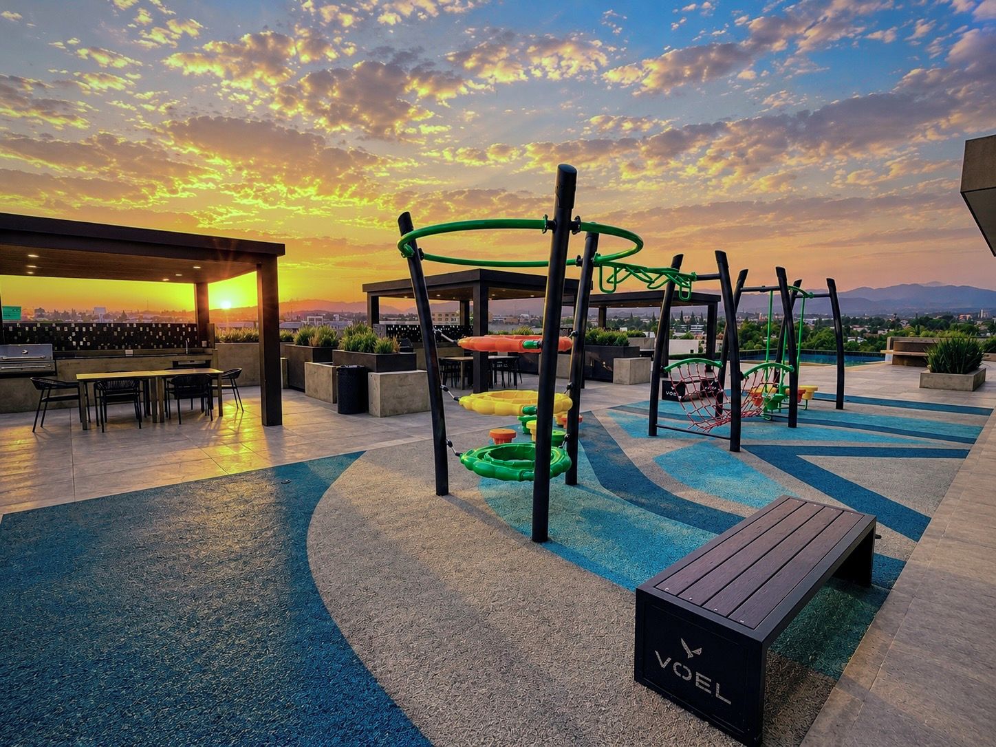 Un espace de jeux extérieur comprenant une balançoire, un toboggan et une structure d’escalade, entouré de sièges et offrant une vue sur le coucher de soleil.