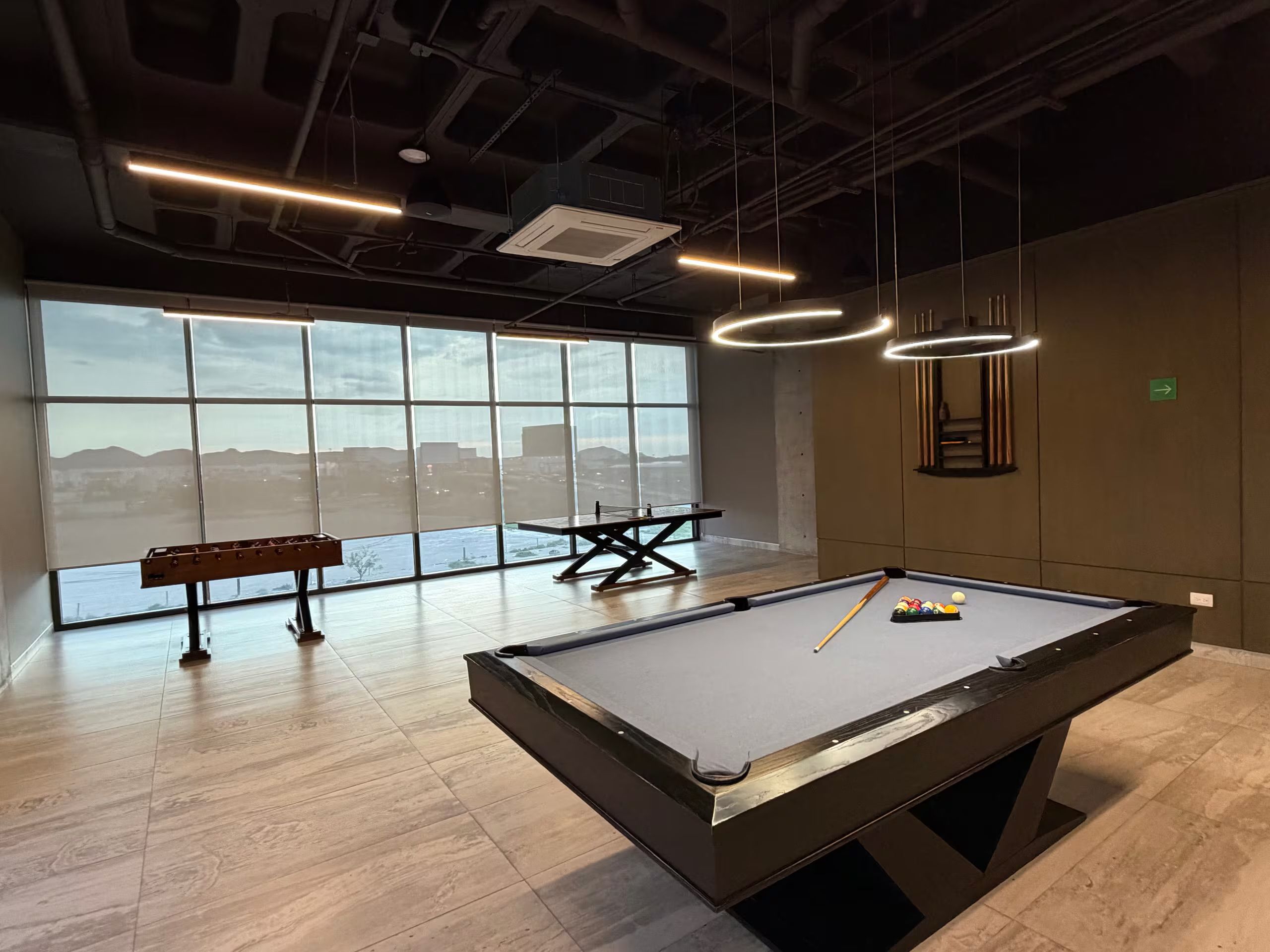 A pool table with a billiard cue and balls, a bench, and a table tennis table in a room with large windows and industrial-style lighting.