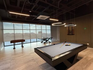 Una mesa de billar con taco y bolas, un banco y una mesa de tenis de mesa en una sala con ventanas amplias e iluminación de estilo industrial.