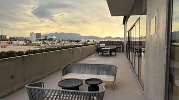 Una terraza en la azotea con mobiliario de mimbre, suelo de hormigón y vistas a un paisaje urbano y montañas a lo lejos.