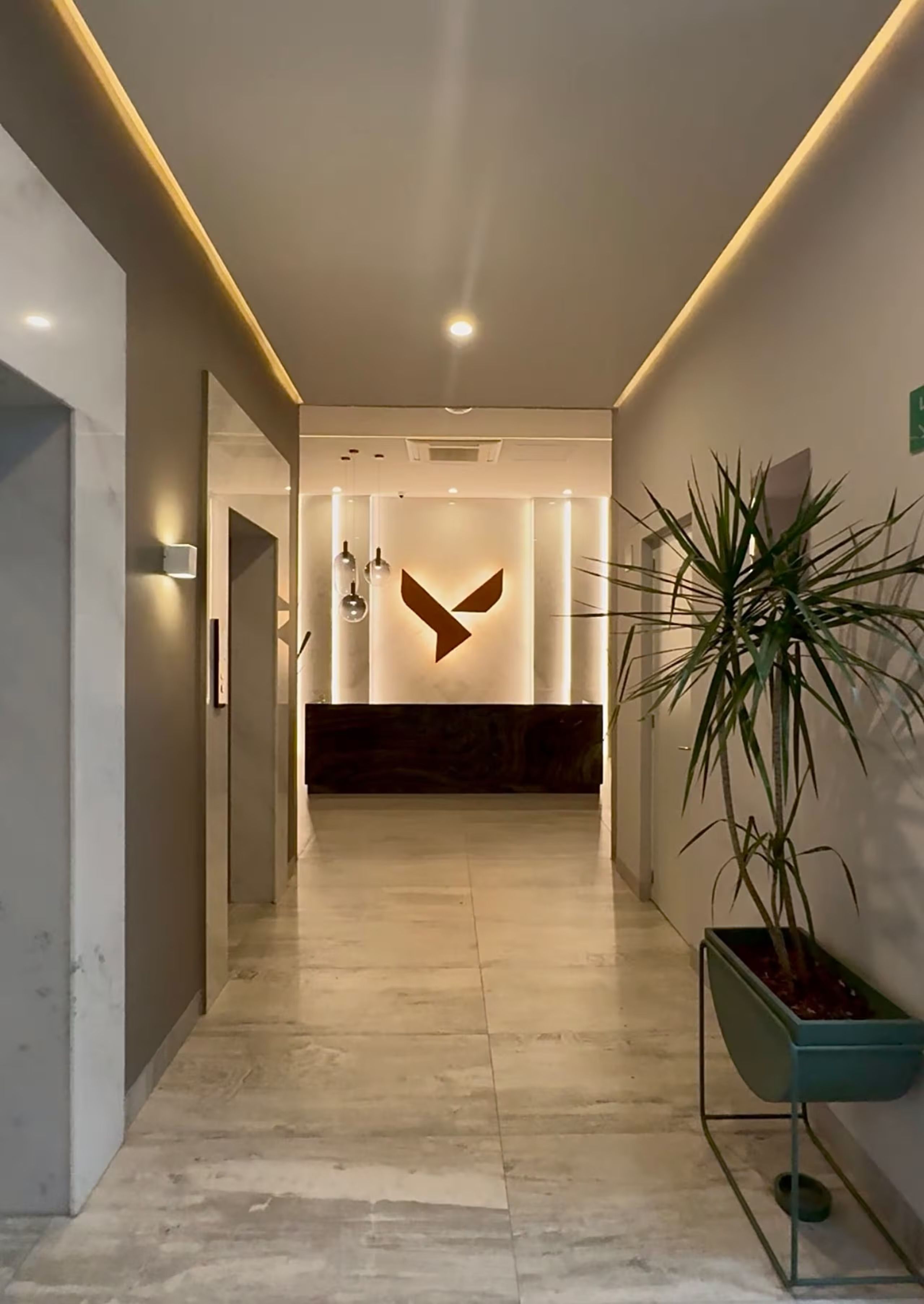 Uma área de recepção moderna com design minimalista, destacando um balcão de atendimento, um vaso de planta e um aparelho de luz fixado na parede.
