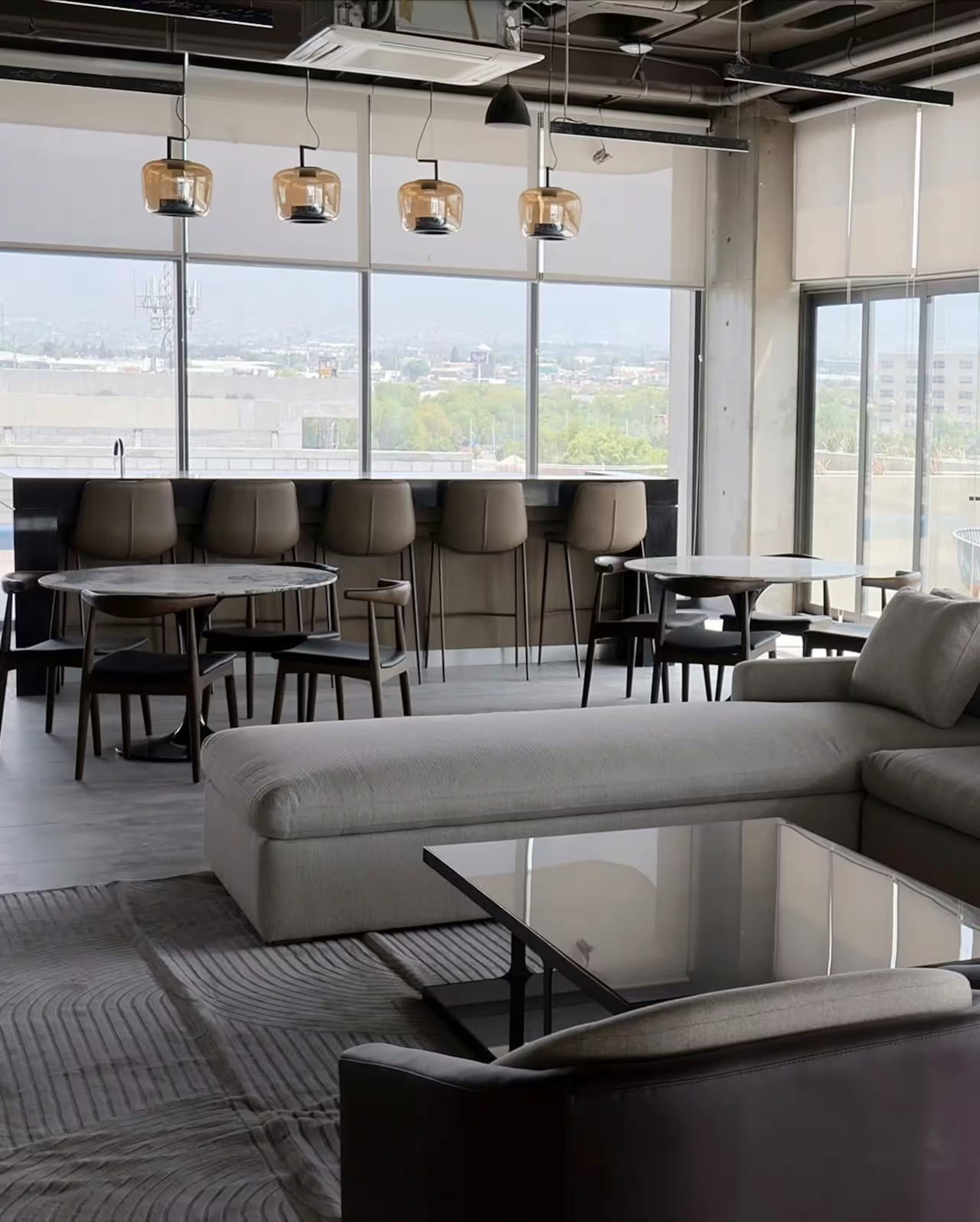 A modern interior with a sectional sofa, round tables, and chairs, large windows with a city view, and pendant lighting.