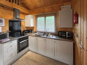 A compact kitchen with wooden walls, a built-in stove, a sink, and a microwave. - Log Cabin Two - UK51960 (Sutton-on-Sea)