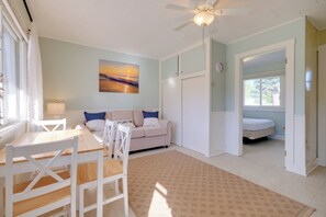 A room with a dining table, chairs, a sofa, a bed, a ceiling fan, a painting, and a window.