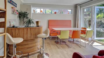 A modern interior with a round wooden reception desk, a white table with orange chairs, and a large window offering a view of greenery.