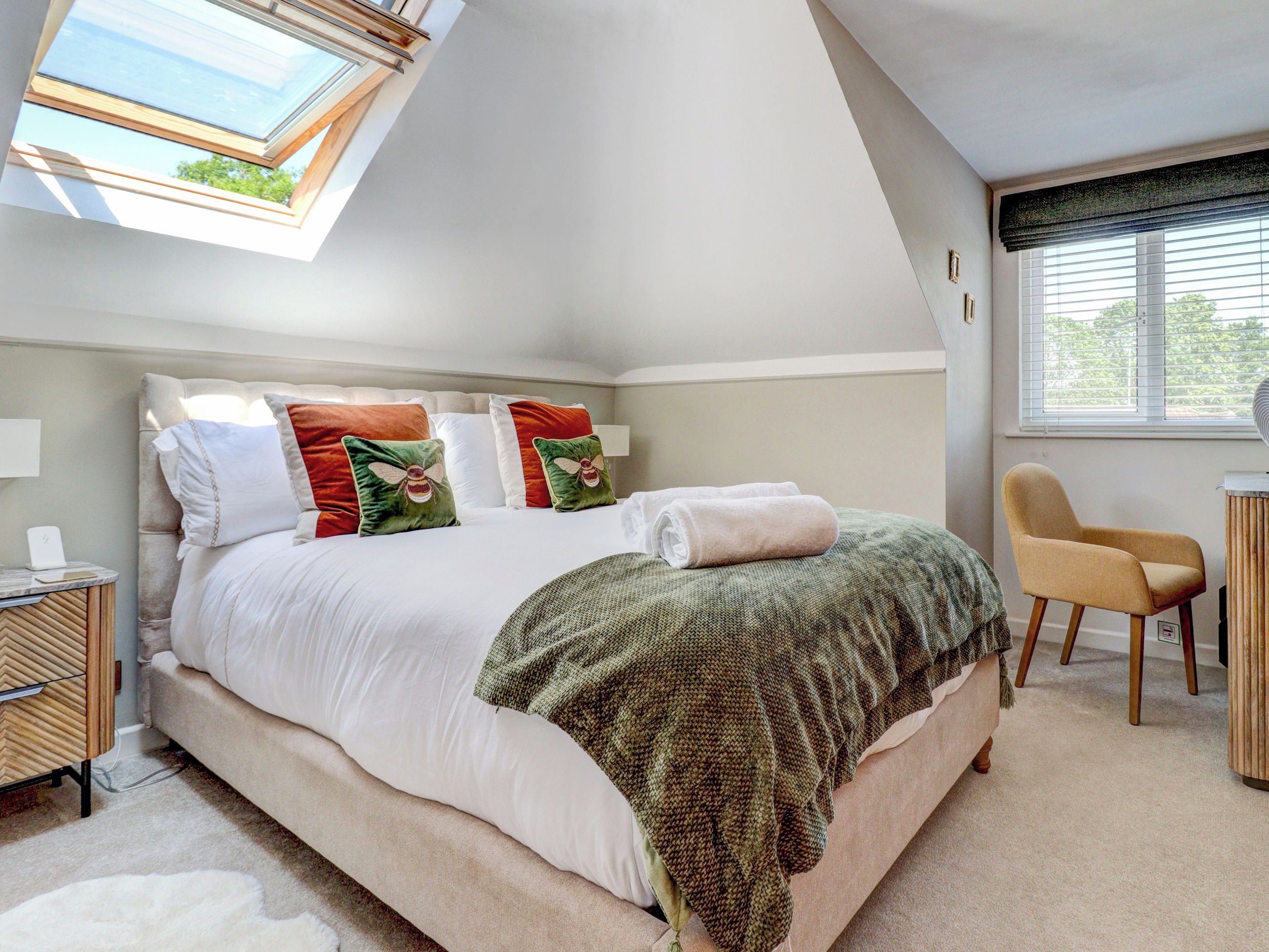 A bedroom with a bed, pillows, a bedside table, a chair, a window, and a skylight.