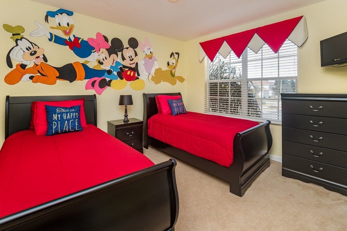 A bedroom with two beds, a dresser, a TV, and a wall mural of Disney characters.