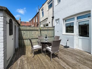 A patio with a round table and wicker chairs, a wooden deck, and a white building with a window. - 2 bedroom accommodation in Guisborough (Guisborough)