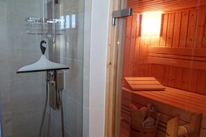 Sala de sauna com chuveiro em compartimento de vidro, bancos de madeira e um banco com cesta.