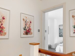 A hallway with framed floral artwork on the walls, a wooden handrail, and a door leading to a bedroom. - The Garden House - UK52180 (Beaworthy)