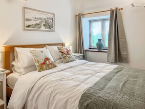 Een opgemaakt bed met witte lakens en decoratieve kussens, een nachtkastje met een lamp, een raam met gordijnen en een lijst met een landschapsfoto aan de muur.