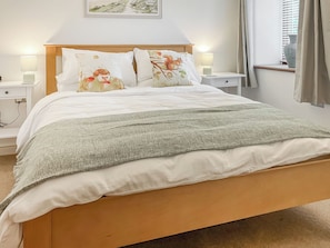 A neatly made bed with a patterned pillow, a bedside table with a lamp, and a window with curtains. - The Garden House - UK52180 (Beaworthy)