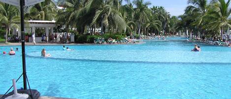 A large outdoor swimming pool surrounded by palm trees, with lounge chairs and people swimming.