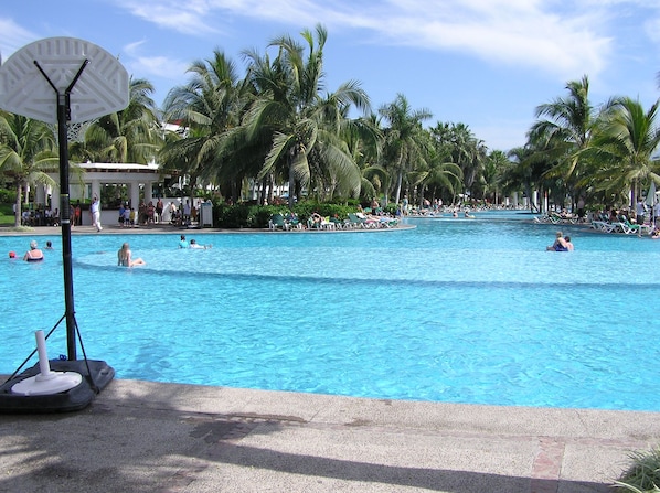 A large outdoor swimming pool surrounded by palm trees, with lounge chairs and people swimming. - PGA golf tournament- 1 bedroom Grand Luxxe villa (Puerto Vallarta)