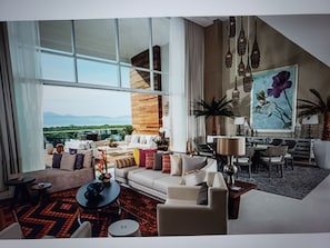 A spacious living area with a large window, a sofa set, a coffee table, and a dining area with chairs. - PGA golf tournament- 1 bedroom Grand Luxxe villa (Puerto Vallarta)