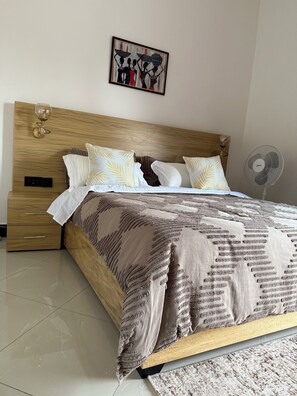 A neatly made bed with a patterned bedspread and pillows, a wooden headboard, a bedside table with a lamp, and a wall-mounted artwork. - The Ember House_Stylish 1BR apartment in kyanja. (Kampala)