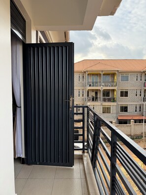 Ein Balkon mit einem schwarzen Metallgitter, gefliestem Boden und Blick auf ein mehrgeschossiges Gebäude mit Balkonen.