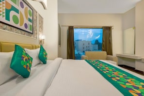 A hotel room with a bed, a sofa, a view of buildings, and a painting on the wall. - Treebo Premium Vedic Stays (Prayagraj)