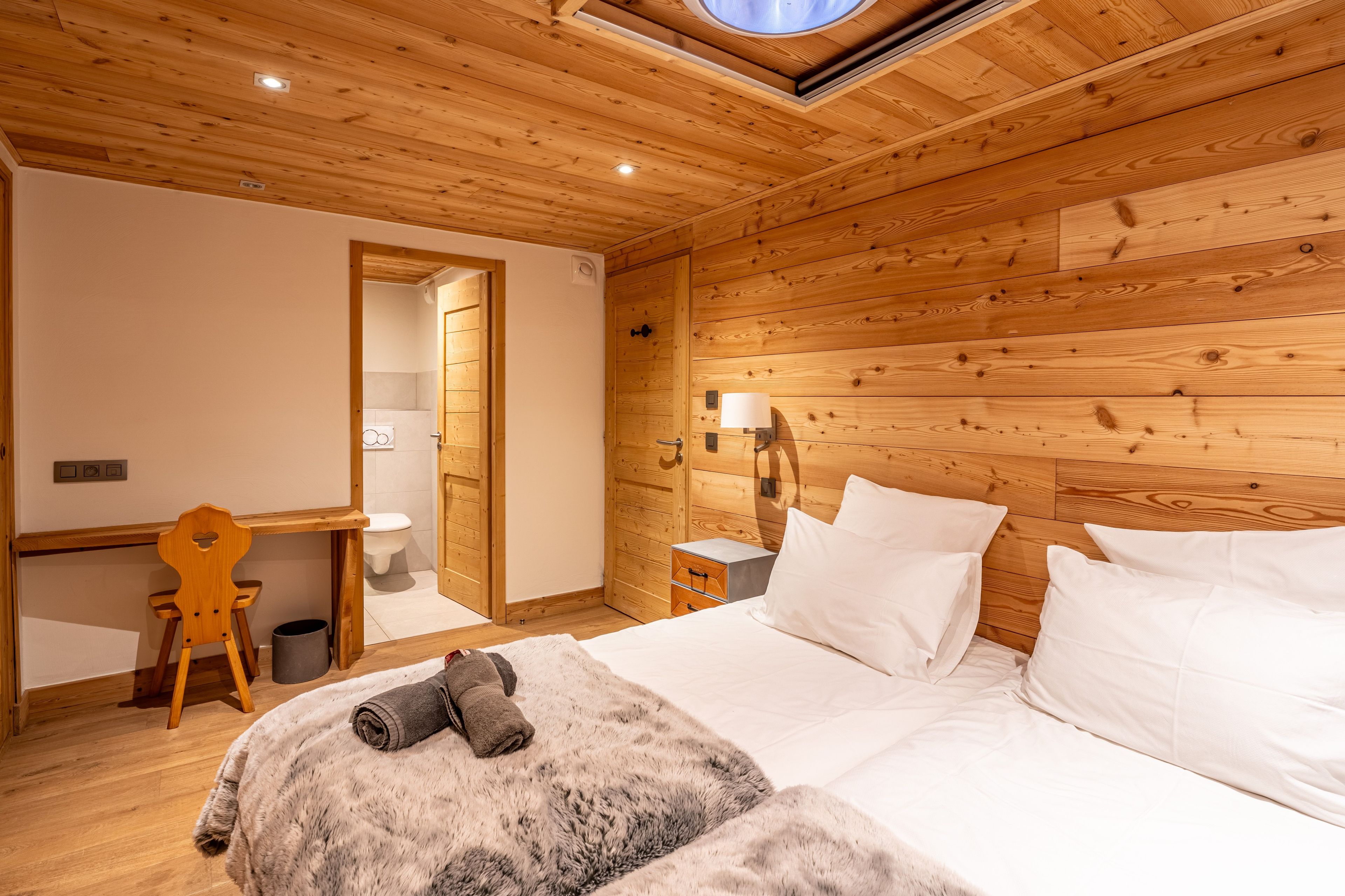 Een knus ingerichte kamer met houten plafond, een bed met witte lakens, een bureau met stoel, en een klein nachtkastje met lamp.