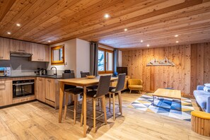 A modern kitchen with wooden cabinets, a dining area with a wooden table and chairs, a sofa, and a patterned rug.