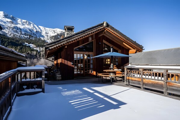 A wooden chalet with a balcony, a table, and a chair, set against a snowy mountain backdrop.