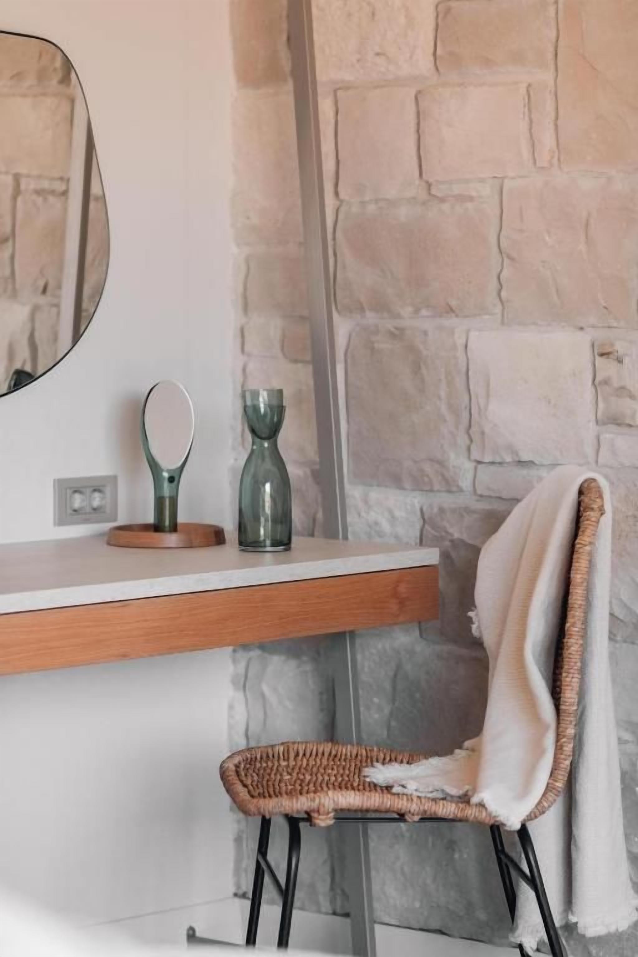 Un baño con pared de piedra, estante de madera con espejo, silla de mimbre y toalla.